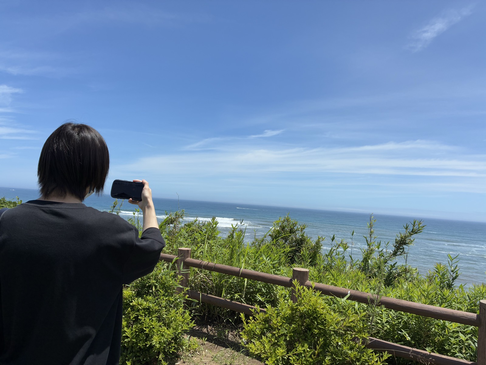 久慈浜海水浴場を見下ろす高台で、スマートフォンを構えて海を撮影する利用者さんの後ろ姿